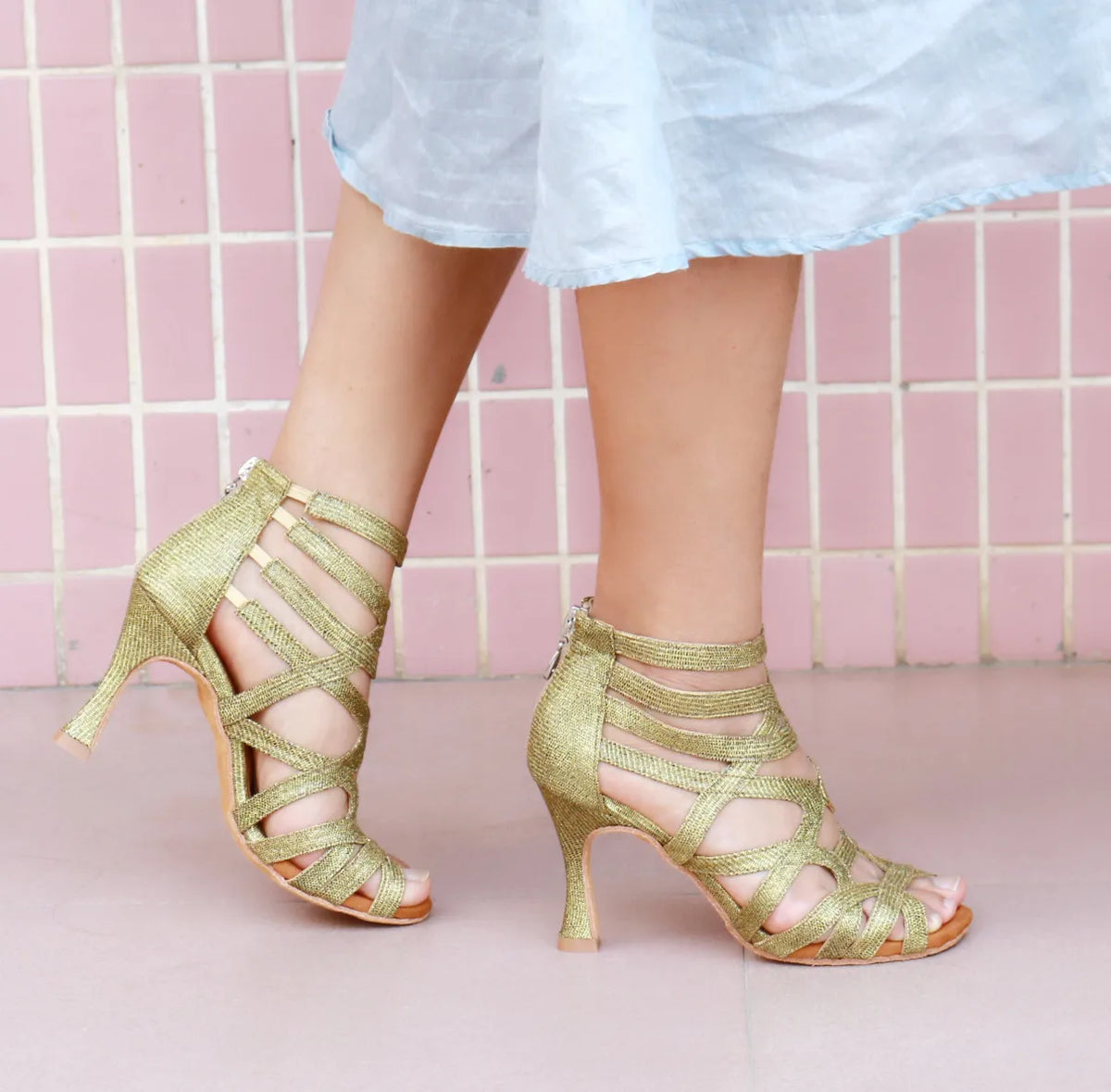 A pair of women's metallic gold strappy high-heeled sandals worn with a light blue skirt, shown against a pink tiled wall background.