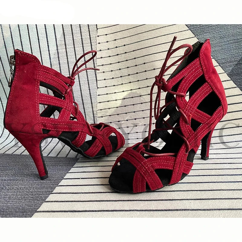 A pair of red suede high-heeled sandals with cutout straps and lace-up front details, displayed on a striped rug with a gray floor background.