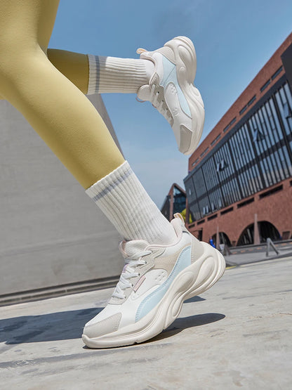 A person wearing chunky white and light blue athletic sneakers with white socks and yellow leggings, captured outdoors on a concrete surface with a modern brick building in the background.