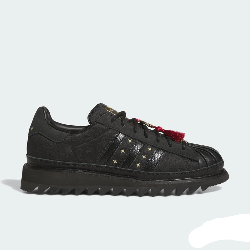 A black low-top sneaker with textured leather panels, gold star accents on the side, a red tassel detail, and a chunky black sole, shown against a light gray background.