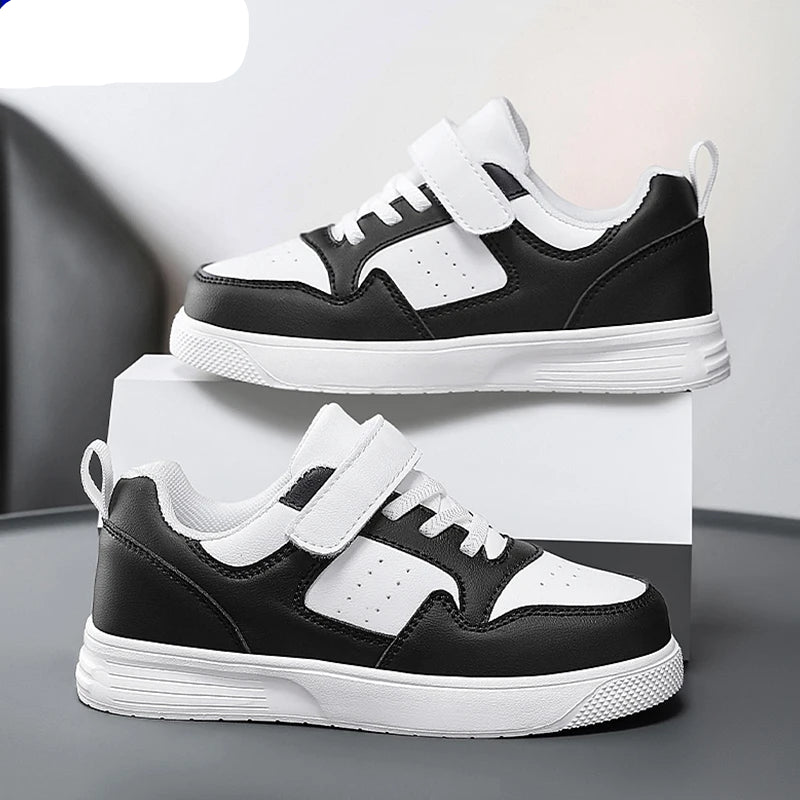 A pair of NBGAGA kids' low-top sneakers in black and white, featuring hook-and-loop straps and thick white soles, displayed on a white block against a gray background.