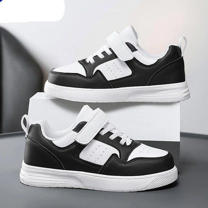 A pair of NBGAGA kids' low-top sneakers in black and white, featuring hook-and-loop straps and thick white soles, displayed on a white block against a gray background.