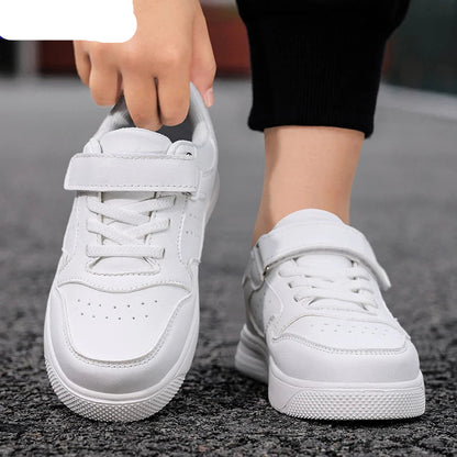 White low-top sneakers with hook-and-loop straps, shown on a person standing on a gray asphalt surface. The person's hand is lifting the strap of the left shoe, and a black knit sock or pant leg is visible.