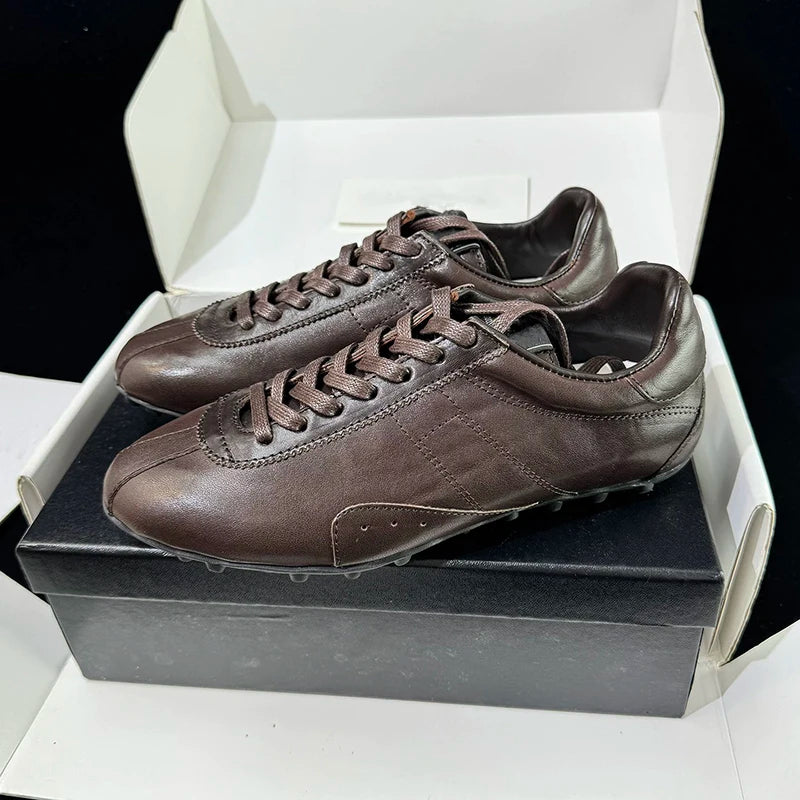 A pair of dark brown leather athletic sneakers resting on a black shoebox inside an open white box, photographed against a black background.