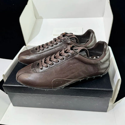 A pair of dark brown leather athletic sneakers resting on a black shoebox inside an open white box, photographed against a black background.