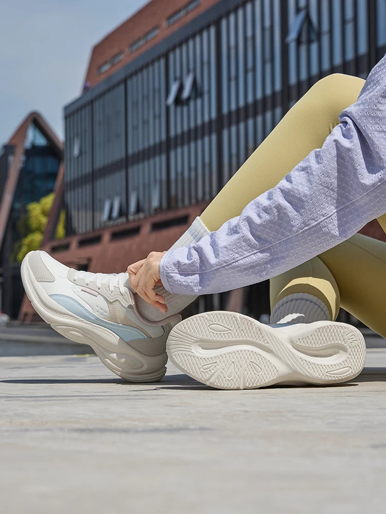 A person sitting outdoors wearing chunky off-white sneakers with light blue accents, paired with pale yellow leggings and a light purple shirt. The background shows a modern building and concrete ground.
