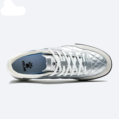 A silver quilted sneaker with white laces, shown from a side profile view on a light gray background. The KELME brand logo is visible on the tongue and inlay of the heel.