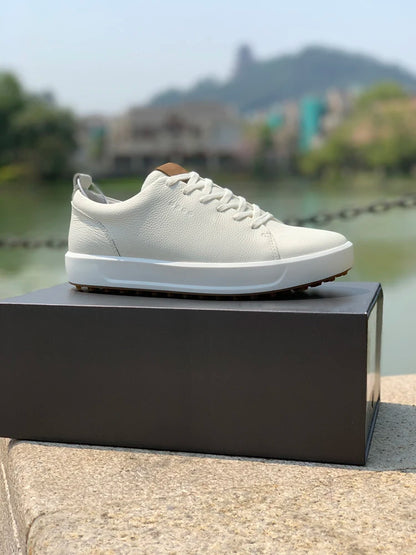 A low-top white leather sneaker with a white sole and laces, resting on a dark gray shoebox outdoors on a stone ledge with a blurred lake and mountain background.