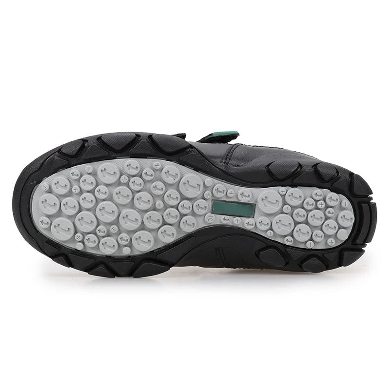 A black UOVO shoe shown from the sole view, featuring a textured black rubber outsole with circular tread patterns and a small green accent tab, on a white background.