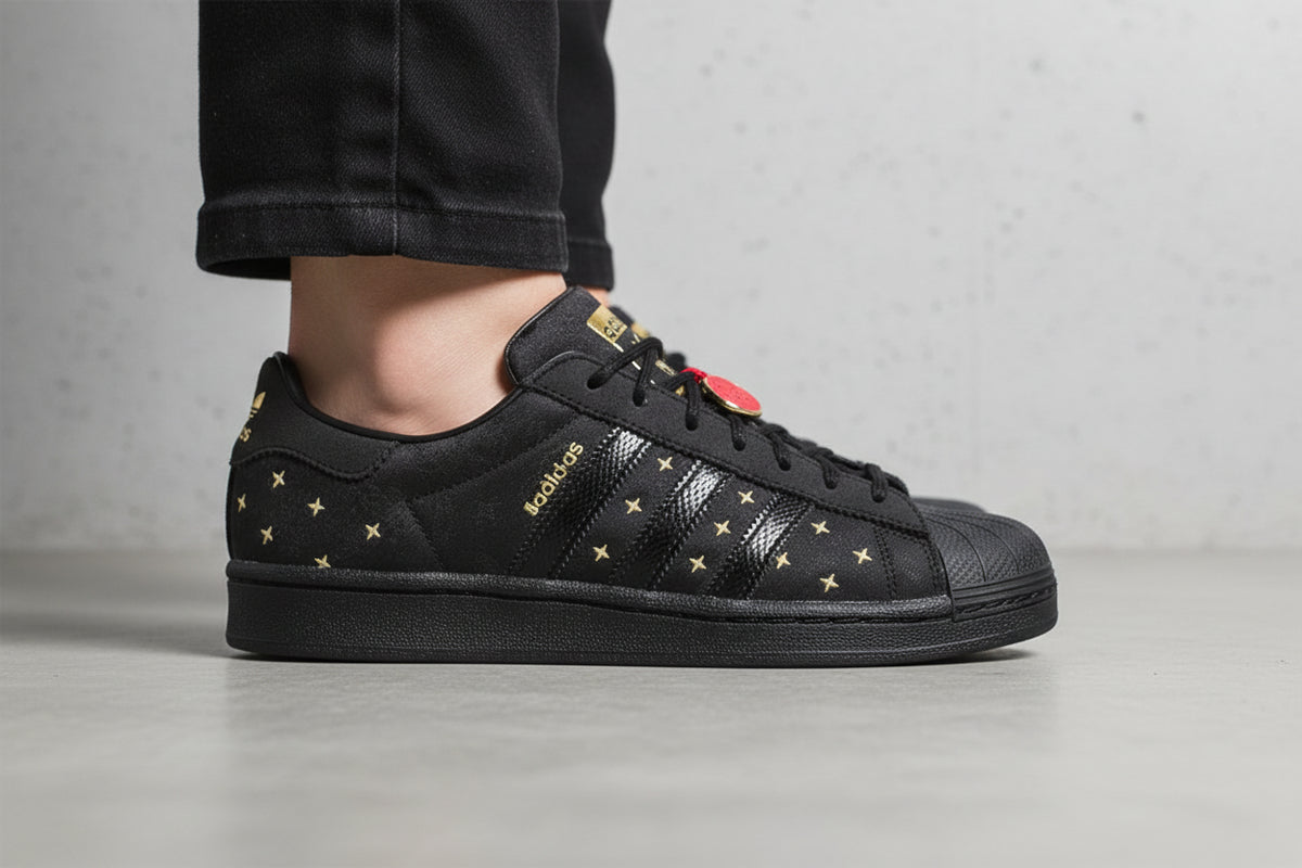 Close-up of a black low-top sneaker featuring textured fabric panels, black laces, gold star accents on the side, and a gold heel tab, set against a light gray background.